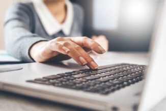 Szary, otworzony laptop. Nad czarną klawiaturą unosi się kobieca ręka