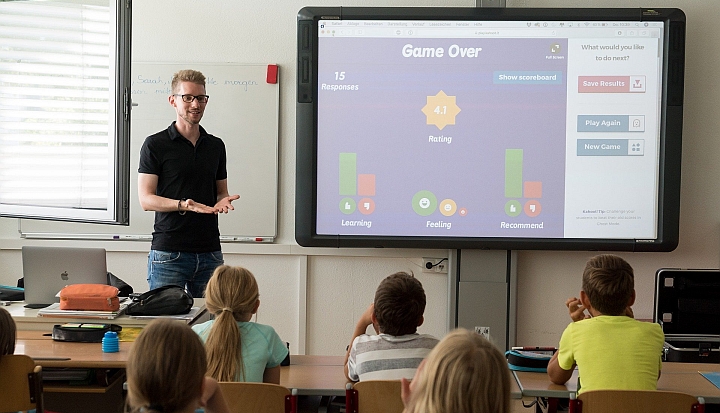 Nauczyciel w czarnej koszulce stoi przodem do uczniów i tłumaczy lekcję, której treść wyświetla się na dużym ekranie