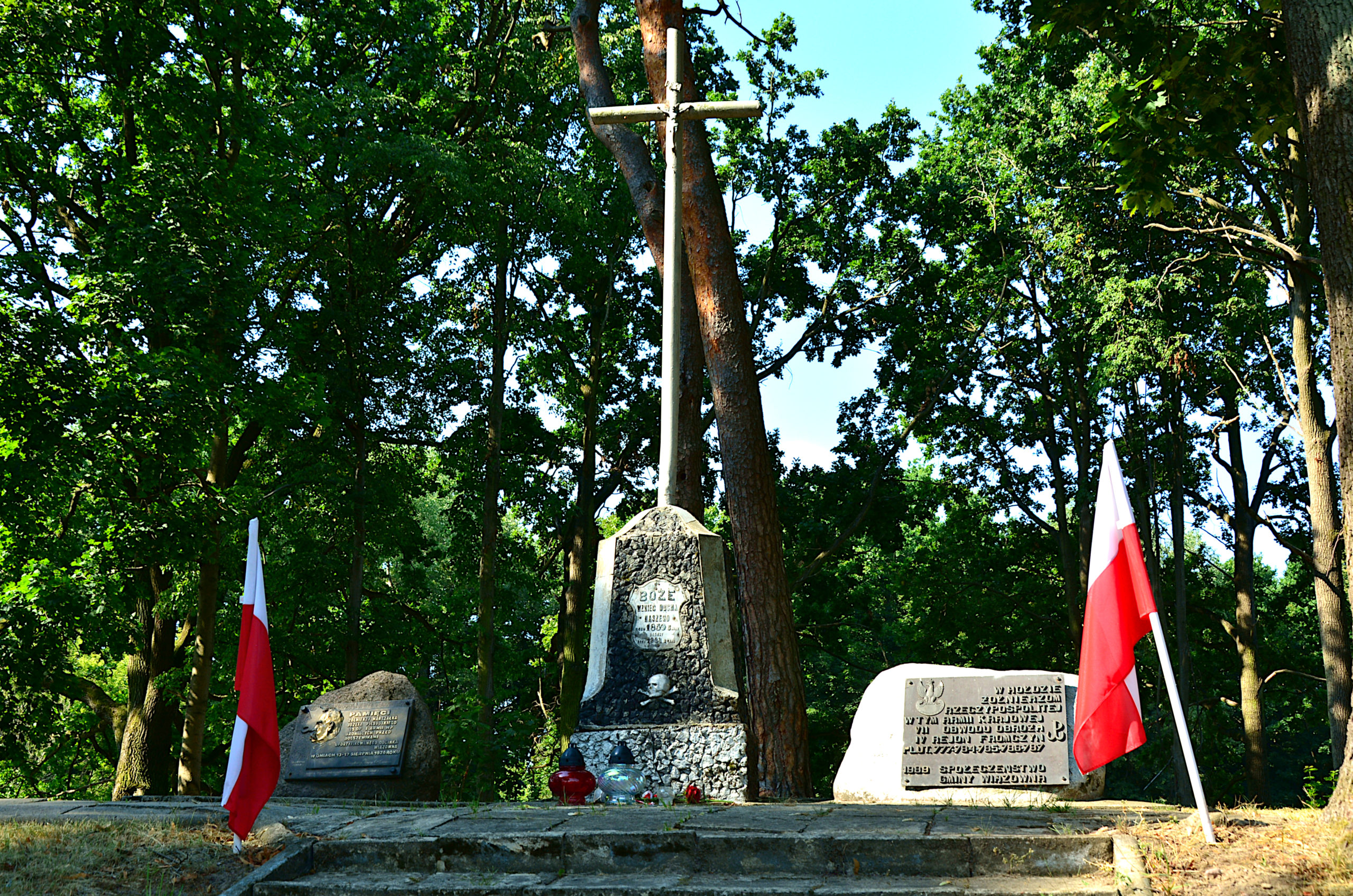 Kopiec w Wiązownie. na środku krzyż, po bokach głazy z tablicami pamiątkowymi. Po obu stronach w ziemię wbito biało-czerowne flagi