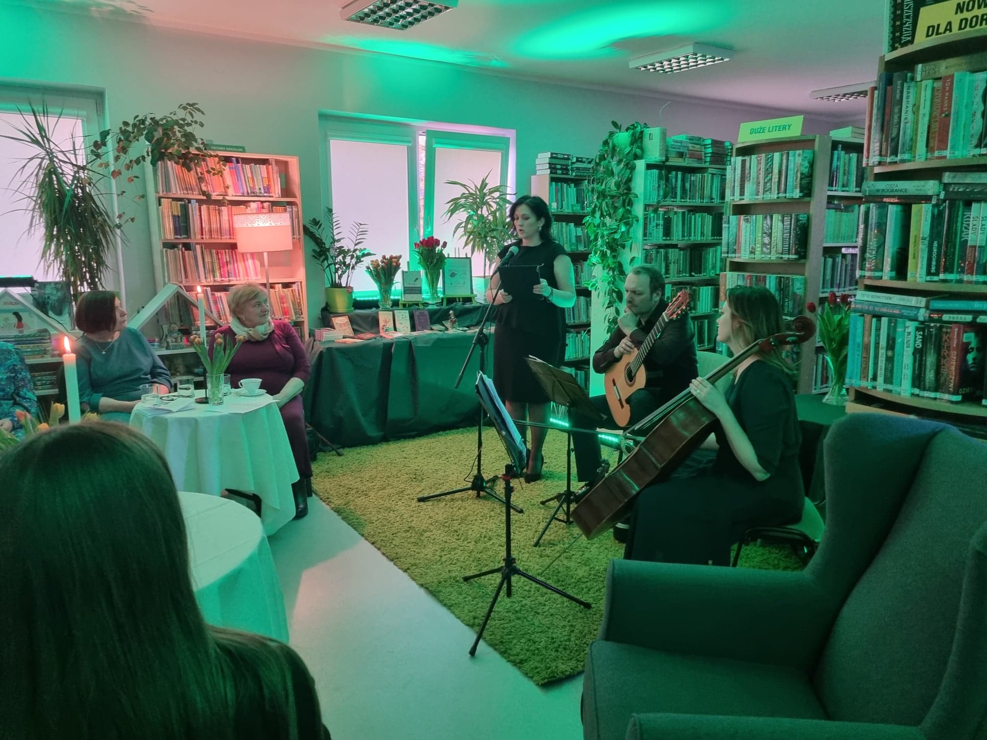 Wieczór poetycko-muzyczny w bibliotece w Wiązownie. Dyrektor biblioteki Marzena Kopka deklamuje wiersz. Z boku siedzi mężczyzna z gitarą na kolanie i kobieta trzymajaca wiolonczelę. W tle regały z książkami.