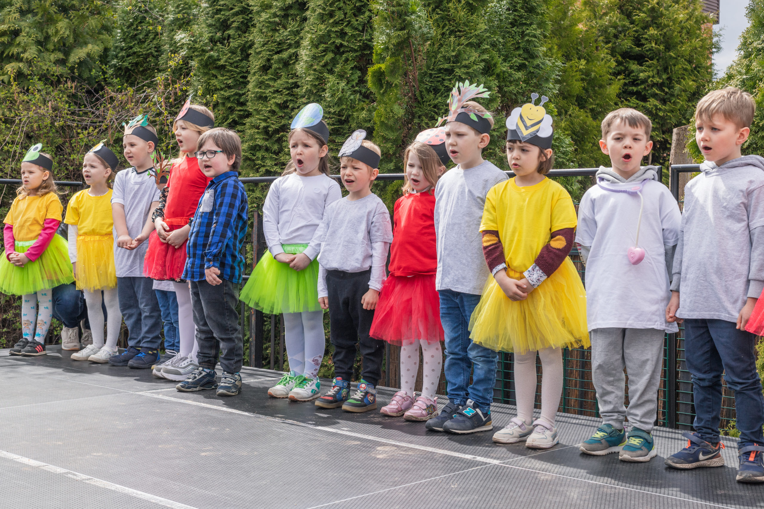 Eko Dzień Sportu w Zakręcie. Dzieci stojące w szeregu śpiewają piosenkę. Mają na głowach opaski z wizerunkami roślin i zwierząt.