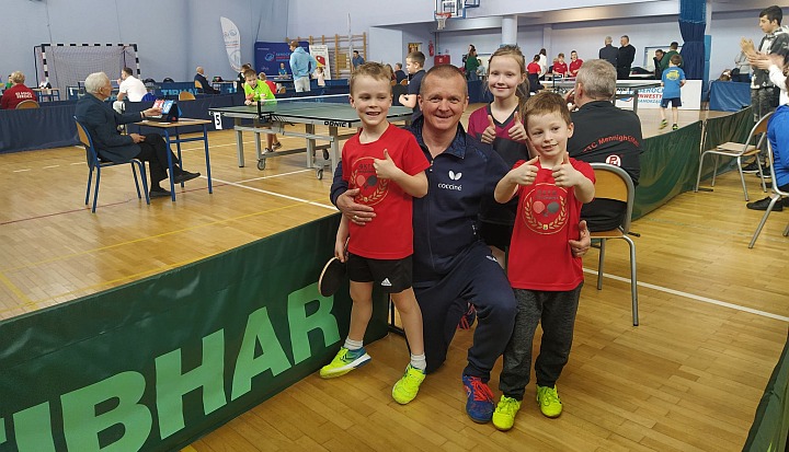 Najmłodsi zawodnicy GKTS Wiązowna rozegrali mecze w tenisa stołowego. Treneromasz Grzybowskiz trójką dzieci w czerwonch koszulkach. Dzieci mają kciuki uniesione w górę.