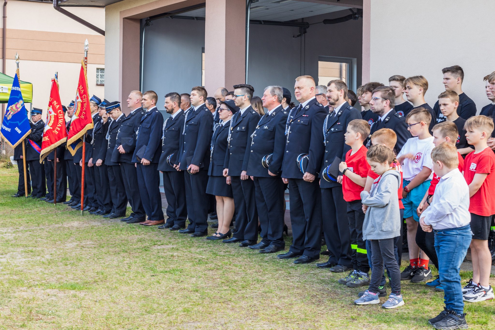 Druhny i druhowie z Ochotniczej Straży Pożarnej oraz młodziki z Malcanowa stoją w galowych mundurach przed strażnicą. S prawej strony stoją dzieci z czerwonych koszulkach.
