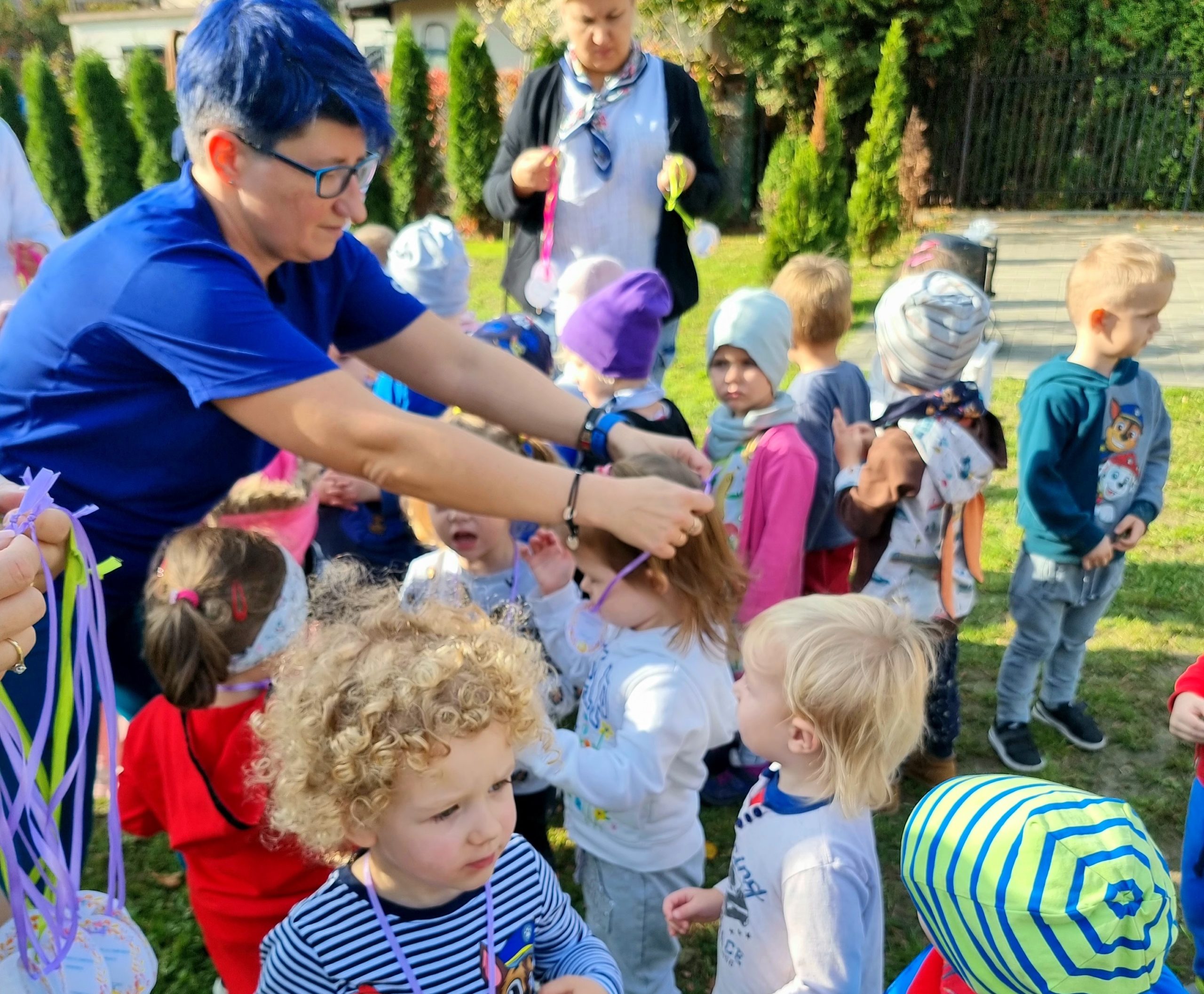 Kobieta z niebieskimim włosami rozdaje medale przedszkolakom za uczestnictwo w olimpiadzie.