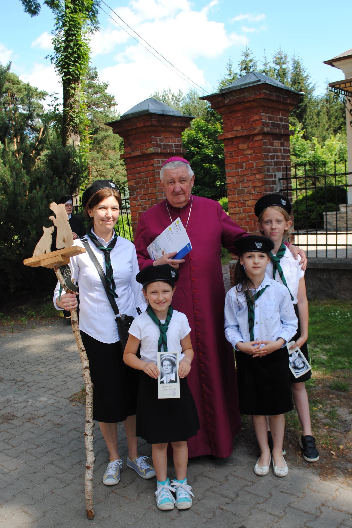 Biskup katolicki z czterema dziewczynkami - skautkami, ubranymi w białe koszule, czarne spódniczki. W tle ceglane ogrodzenie i zieleń.