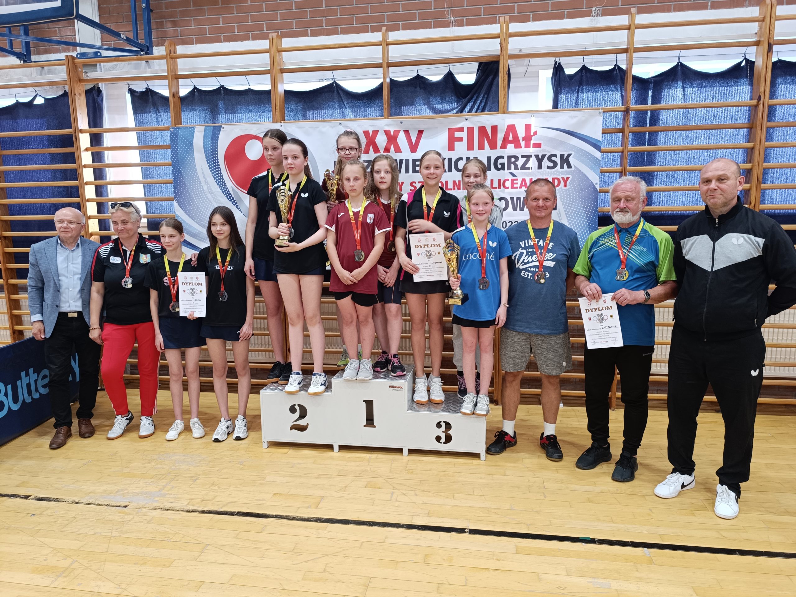 Najlepsze drużyny na Mazowszu w tenisie stołowym dziewcząt drużyny szkolne stoją na podium dla zwycięzców. W tle drabinki gimnastyczne.
