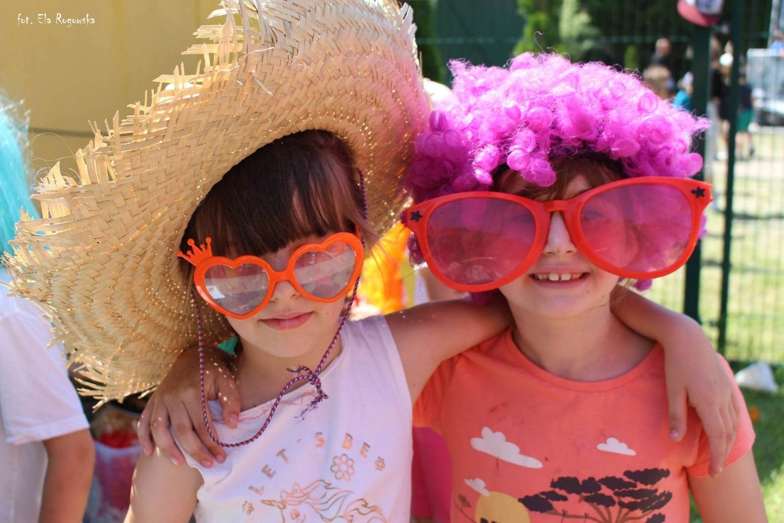 Dwie dziewczynki w okularach, ubrane w kapelusz i perukę czekają na zdjęcie z fotbudki.