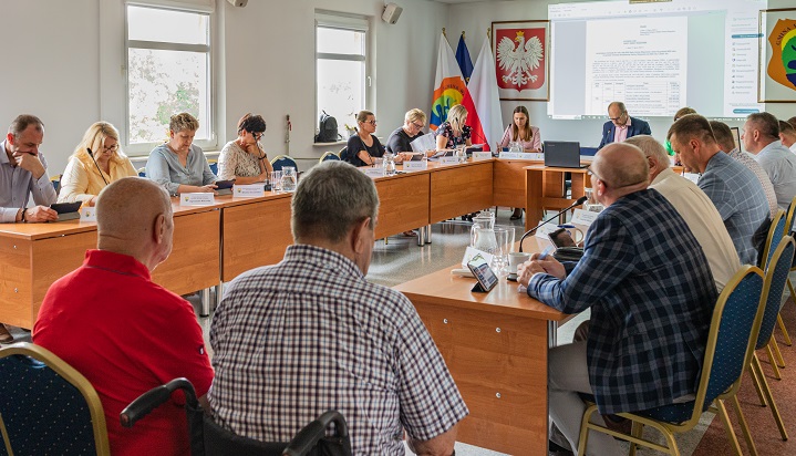 Nadzwyczajna sesja Rady Gminy Wiązowna 17.07.2023 r. Radni siedzą przy stołach ustawionych w literę u. Na ścianie wyświetla się plik z uchwałą. Na ścianie godło Polski i herb gminy.