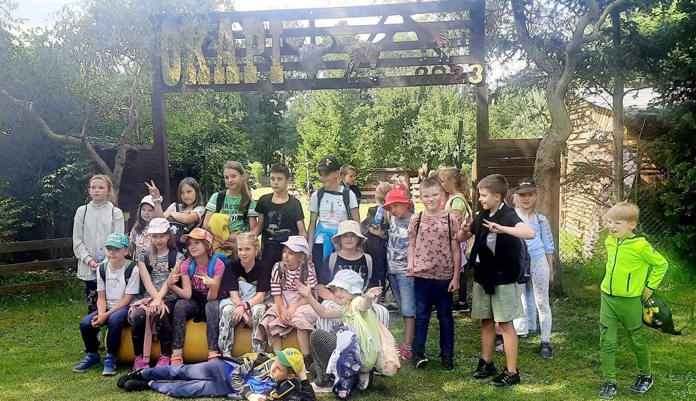 Wakacje blisko domu w szkole w Gliniance. Duża grupa dzieci w letnich strojach [ozuje do wspólnego zdjęcia. W tle napis Okapi i zieleń.