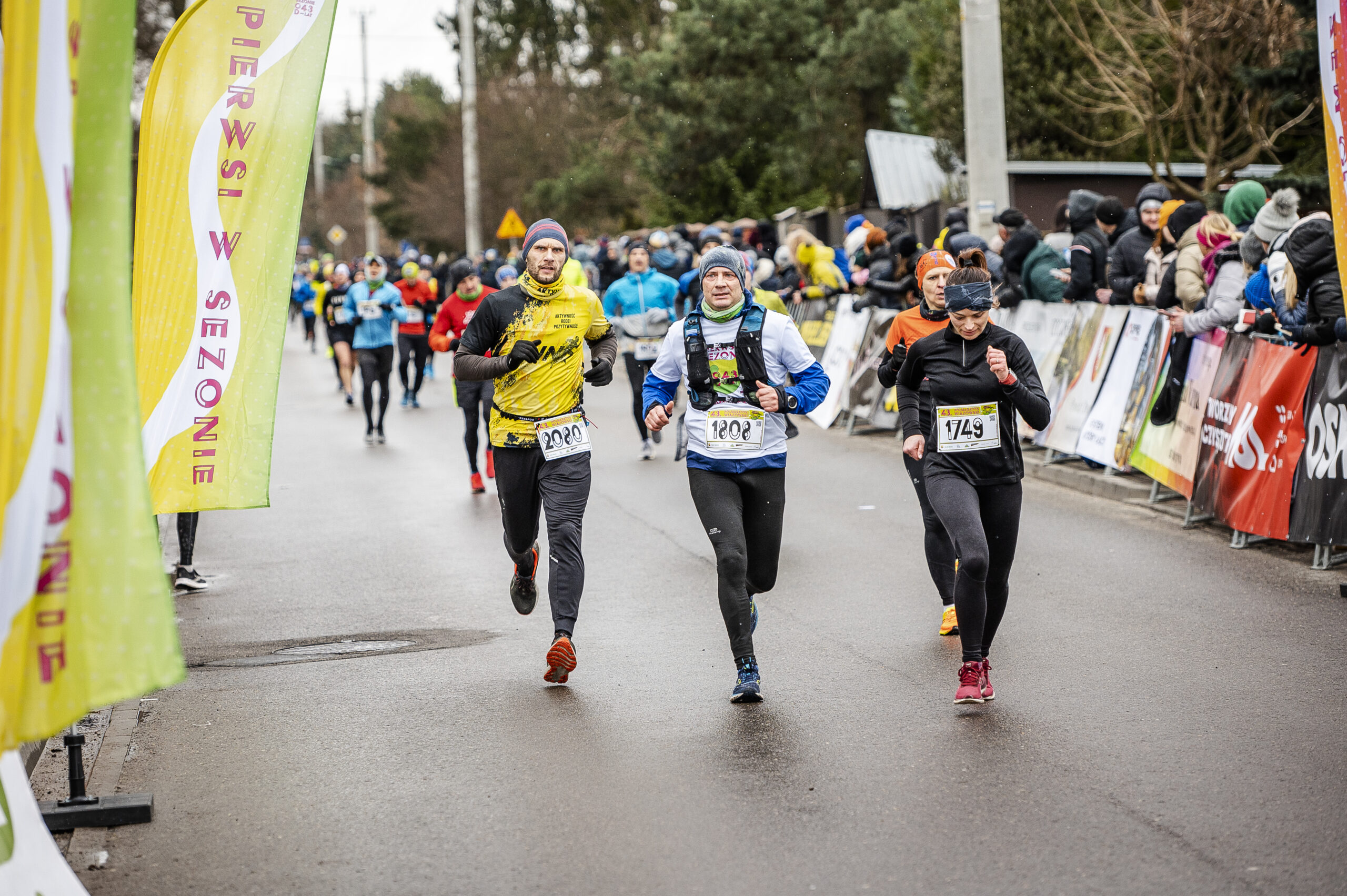 Trzech biegnących mężczyzn. Za nimi kolejni biegacze. Z lewej strony banery żółto-białe. Z prawej za barierkami stoją dorośli i kibicują biegaczom.