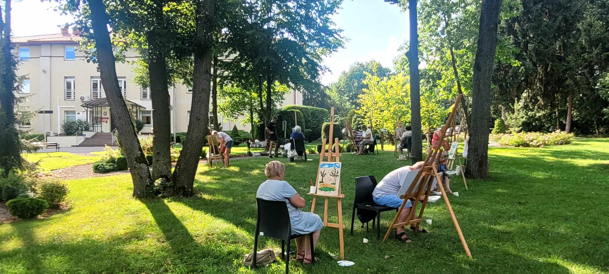 Teren zielony wśród drzew. Na trawie stoją sztalugi, przed nimi siedzą osoby i malują.