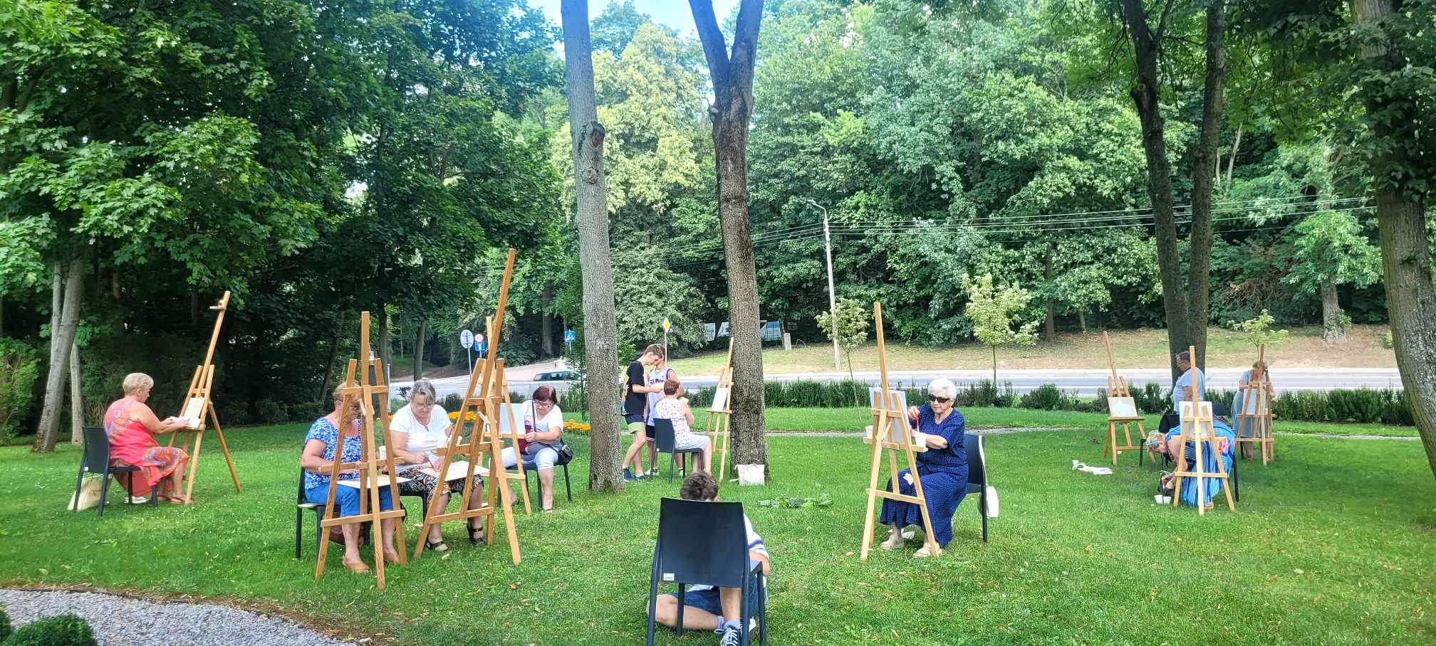 Teren zielony. Drzewa. Na trawie stoją sztalugi, przed nimi siedzą osoby i malują.