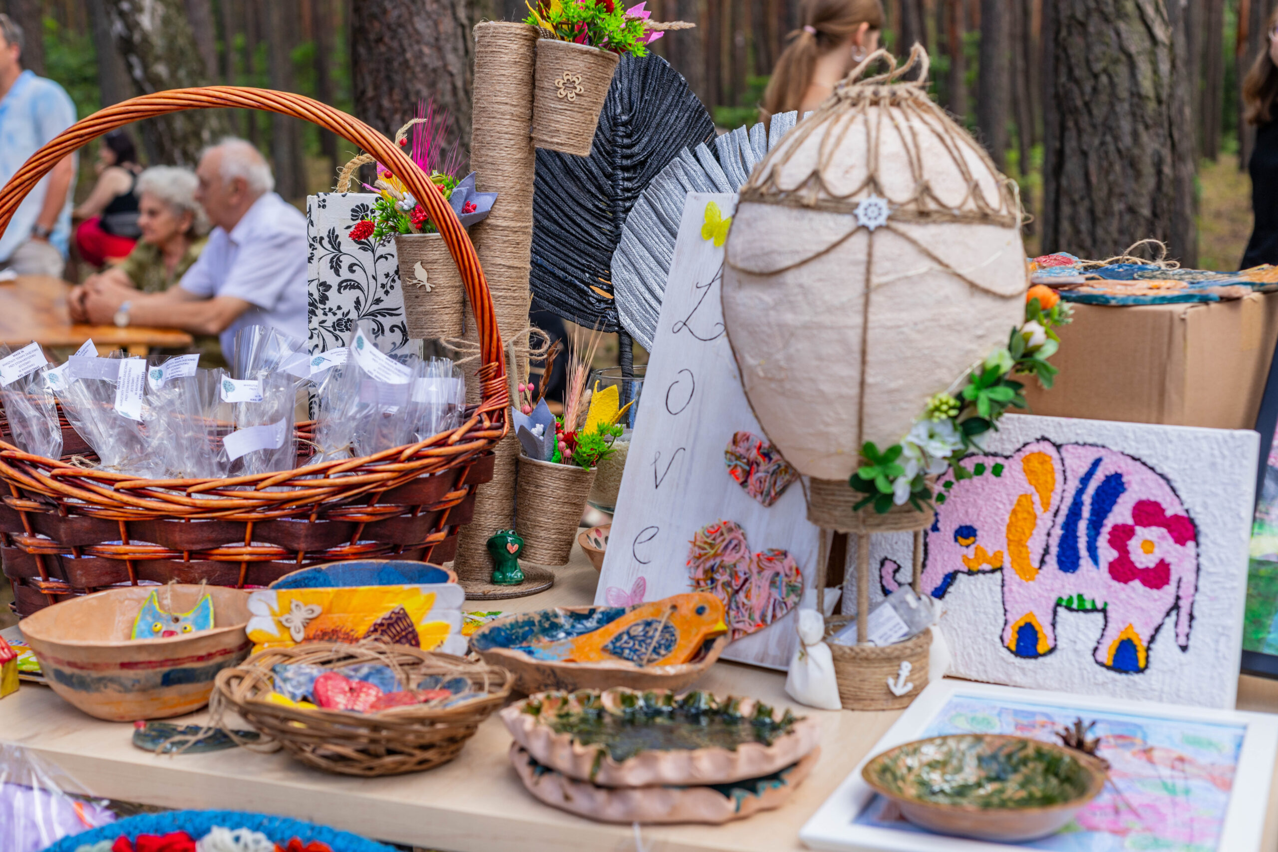 Stół z rękodziełem, a na nim misy i kolorowe ptaki oraz koty z gliny, wiklinowy koszyk, wyklejanka - słoń, obrazek w białej ramce.