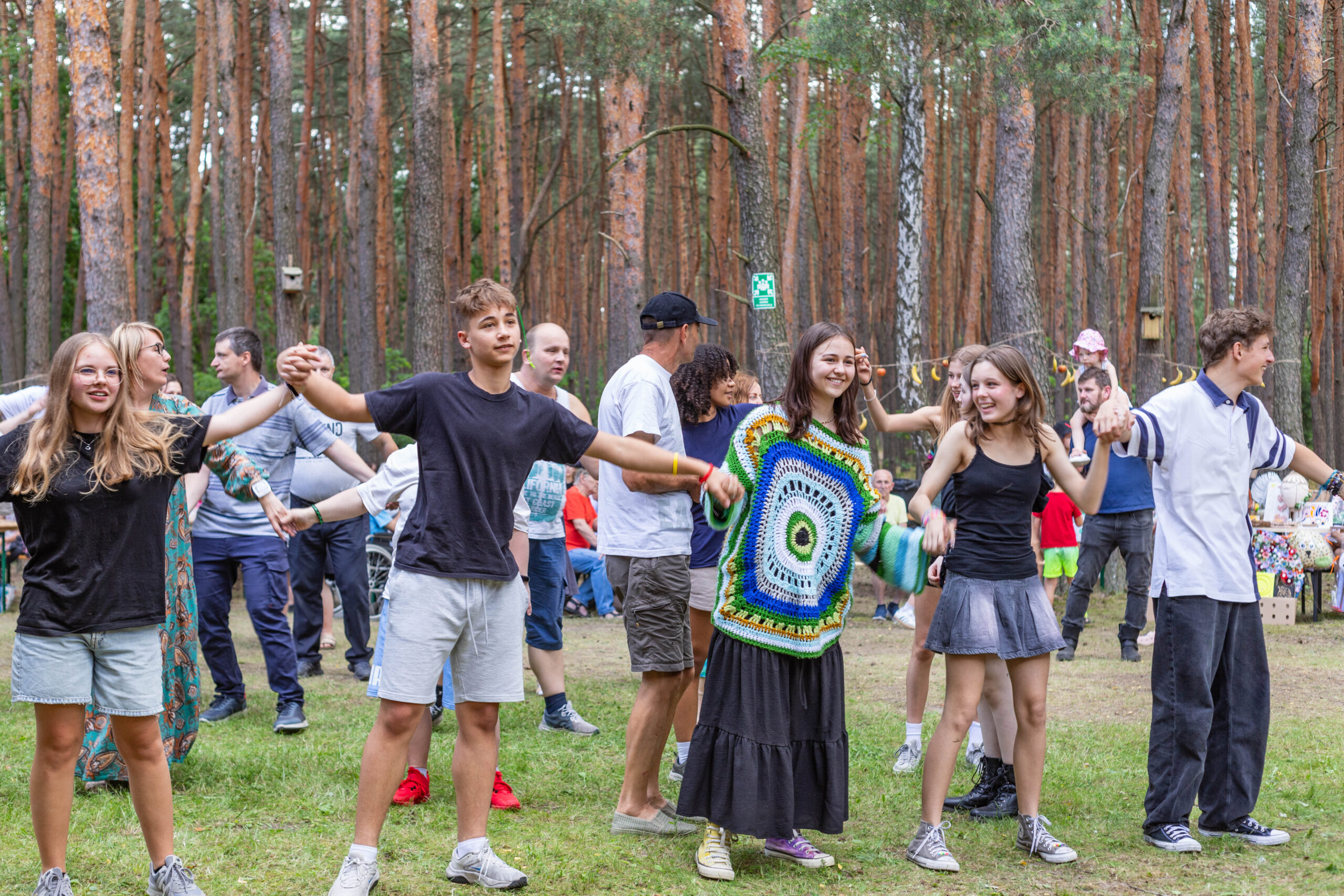 Grupa kolorowo ubranych i roześmianych młodych ludzi trzyma się za ręce. W tle drzewa.